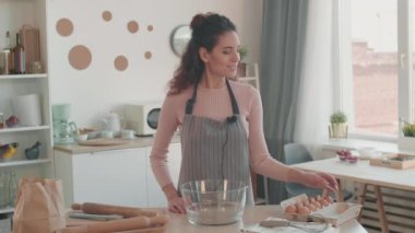 Kahverengi saçlı, önlük takan, mutfak masasının yanında duran, kamerada yemek pişirmek için gerekli olan aletleri gösteren ve tanımlayan orta karar beyaz kadın portresi.