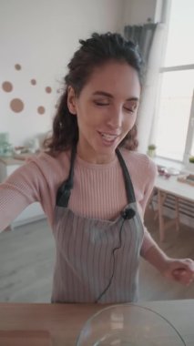 Dikey el bilgisayarı, genç beyaz bir kadının mutfak video blogu için onu çekerken çekilmiş görüntüsü, çırpma, yumurta, oklava, konuşma, gülümseme, kameraya el sallama.