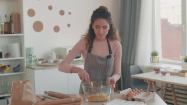 Orta boy beyaz kadın nüfuzlu, mikrofonlu önlük takmış mutfak masasında durmuş, büyük cam kasede yumurta çırpıyor, konuşuyor ve gülümsüyor.