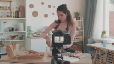Kahverengi saçlı beyaz kadın vlogger 'ın sol orta boy fotoğrafını çekerek mutfak masasında duruyor, hamur ile kağıt torbadan cam kaseye un ekliyor, konuşuyor, önündeki kameraya bakıyor.