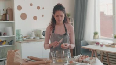 Üzerinde mikrofon olan önlük giyen, mutfak masasındaki büyük cam kaseye yumurtalarını kıran, konuşan, gülümseyen ve kameraya bakan beyaz bir kadının orta ölçekli zaman çizelgesi.