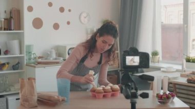 Orta boy beyaz kadın video blogcusu mutfakta video ekipmanlarıyla duruyor, krem şantiyle muffinleri süslüyor, gülümsüyor, kamerada konuşuyor.