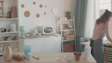 Beyaz önlüklü kadın blogcunun orta ölçekli görüntüsü mutfakta dikilirken, fırında kek yapmak için yürürken, koklarken, onları gösterirken, kamerada konuşurken ve konuşurken.