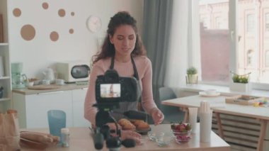 Orta boy beyaz kadın video blogcusu mutfak masasının yanında çöreklerle duruyor, kamerada pişirme süslemeleri olan küçük cam kaseler gösteriyor, tripodun önünde duruyor.