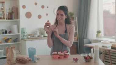 Kıvırcık saçlı, önlük giyen, mutfakta duran, gülümseyen, kamerada konuşan, üzerinde krem şanti ve böğürtlen olan çörekleri tutan orta boy beyaz kadın nüfuzu.