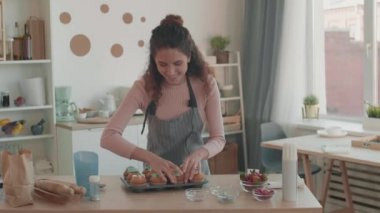 Mutfak masasının yanında duran önlüklü, beyaz tenli, genç bir kadın etkisinde orta boy POV. Kadın kek kağıdından kek alıyor, bunu gösteriyor, konuşuyor, kameraya gülümsüyor.