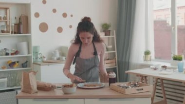 Orta karar beyaz bir kadın mutfak masasında duruyor, ev yapımı kurabiyeleri ikiye bölüyor, kokluyor ve tadıyor, gülümsüyor, kamerada konuşuyor.