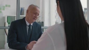 Kafkas erkek İK müdürünün kağıt okurken orta boy yakın plan fotoğrafı. Ofiste onun karşısında oturan uzun saçlı kadın adayla konuşuyor.