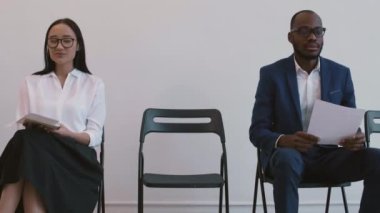 Orta uzunlukta Asyalı genç kadın ve Afrikalı erkek sandalyelerde oturuyor, gülümsüyor, sohbet ediyor, beyaz bir erkek başvuru yaptıktan sonra onlara katılıyor.