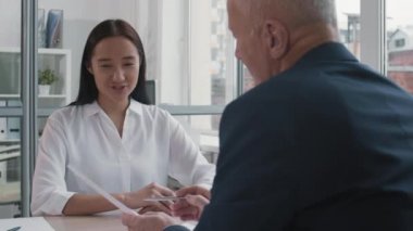 Beyaz tişörtlü, gülümseyen, konuşan, tanınmayan gümüş saçlı İK yöneticisinin önünde masa başında oturan Asyalı genç bir kadın adayın göğsü yukarıda.