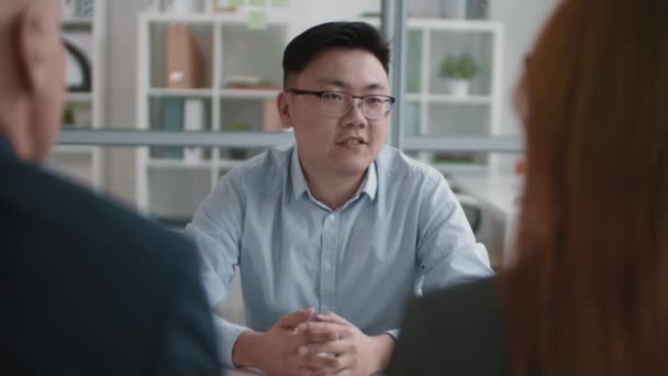 オフィスのデスクに座っている赤い髪のマネージャーと男性の同僚の胸の上の肩 若い自信のあるアジア人男性に直面して面接で質問に答える — ストック動画