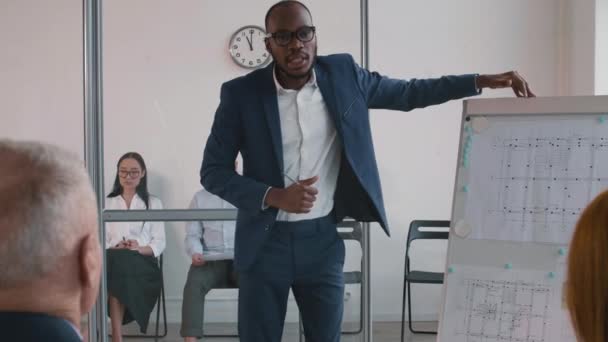 身穿蓝色西服的中等个子的非洲青年商人站在白板旁边 上面有图表 为同事们做演讲 两个人在玻璃墙后面的房间里等着 — 图库视频影像