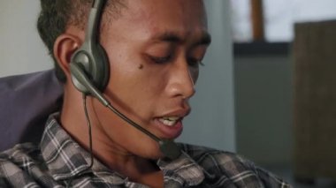 Kareli gömlek ve kulaklık takmış genç Asyalı yardım masası çalışanı koltuğa oturur, müşteriye telefonla danışır ve not defterine notlar yazar.