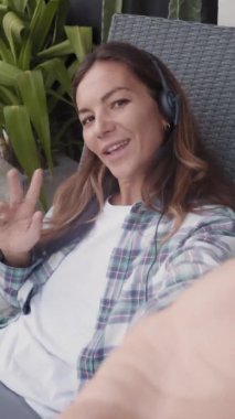 Dikey görüntüde, kadın serbest çalışan, kulaklık takıyor, hasır sandalyede dizlerinin üstünde dizüstü bilgisayarla oturuyor, elinde kamerayla video konuşması yapıyor.