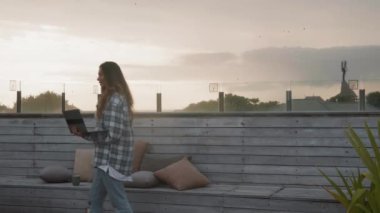 Genç ve güzel bir kadının gündelik giysiler ve kulaklıklar takıp çatıdaki ahşap terasta dizüstü bilgisayarını tutarken ve video bağlantısıyla arkadaşıyla konuşurken orta ölçekli bir fotoğrafı.