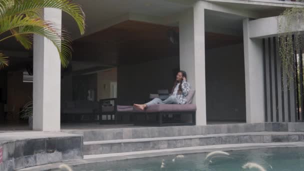 Wide shot of young beautiful woman wearing casual clothes and headset sitting on sofa near pool in modern house with laptop on her knees and communicating with customer by telephone — Stock Footage Wide Shot Young Beautiful Woman Wearing Casual Clothes Headset Sitting Royalty Free Stock Footage