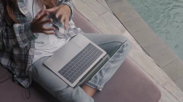 穿着休闲服 牛仔裤和耳机坐在游泳池旁边沙发上 膝上型笔记本电脑 并通过视频链接与同事交谈的高视镜 — 图库视频影像