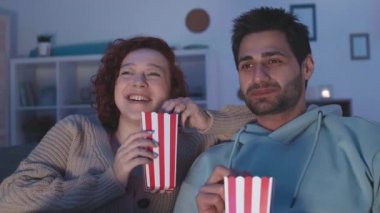 Koyu saçlı, melez, beyaz bir kadının yanında oturan genç adamın yakın plan fotoğrafı. İnsanlar çizgili kırmızı ve beyaz karton kutulardan patlamış mısır yiyor, gülüyor, evde televizyon izliyorlar.