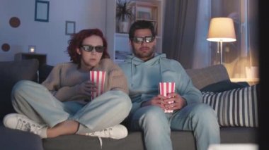 Karışık ırktan genç bir adamın evdeki kanepede oturan, elinde patlamış mısır kutularıyla oturan, 3D gözlük takan, korkan, geri atlayan ve üzerine patlamış mısır serpiştiren kızıl saçlı bir kadının duruşu.