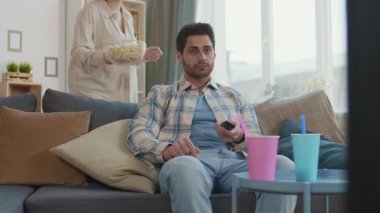 Orta uzunluktaki melez genç adam kanepede oturuyor, uzaktan kumandalı, kızıl saçlı beyaz kız arkadaşıyla televizyon kanallarını değiştiriyor patlamış mısırlı kase getiriyor, ona sarılıyor, konuşuyor.