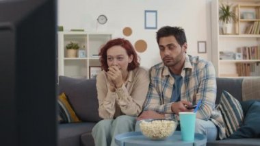 Orta uzunlukta, kızıl saçlı beyaz kadın ve melez adamın evdeki kanepede oturup film izlemesi, korkması.