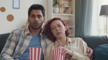 Oturma odasındaki koltukta oturan orta boy duygusal beyaz kız, siyah saçlı melez erkek arkadaşı ona sarılıyor. Elinde patlamış mısırla karton kutu tutan çift filmden korkuyor.