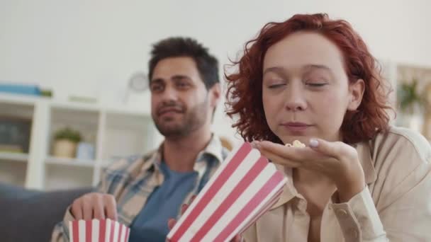 赤髪の白人の女の子のクローズアップとぼやけた混戦の男は家に座って 赤と白の縞模様の紙の箱からポップコーンを食べて 興味深い映画を見て — ストック動画