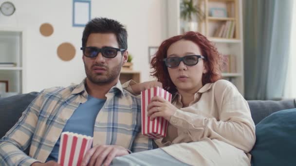 若い混合人種のボーイフレンドと一緒にソファに座っている生姜髪の白人女性の中出し カップルは3Dメガネをかけ 映画を見て 怖がって ポップコーンを散乱 — ストック動画