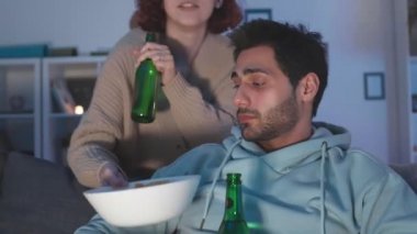 Genç melez adamın evdeki kanepede oturduğu yakın plan, kızıl saçlı, beyaz kız arkadaşı ona atıştırmalık ve yanaktan öpücükler getiriyor. Televizyon izleyen ve bira içen çiftler.