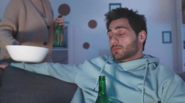 Evdeki koltukta oturan, elinde bira şişesi olan, televizyon izleyen, kızıl saçlı, beyaz kız arkadaşının ona atıştırmalık getirdiği ve yanağından öptüğü genç bir adamın yakın çekimi.