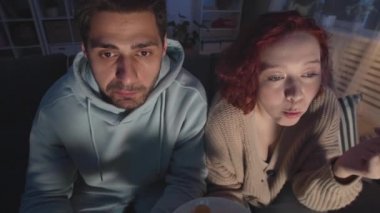 Yüksek açılı orta boy, beyaz ırktan bir adam ve beyaz bir kızın dairede oturup cips yerken, şişe bira içerken, sohbet ederken, geceleri görünmez TV izlerken.