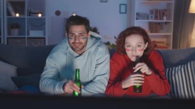 Kırmızılı, mavi elbiseli, yüzü çizgili beyaz bir adamın, gece televizyonda maç izlerken favori takımı için tezahürat yaparken, şişe bira içerken orta boy beyaz bir kadın resmi.