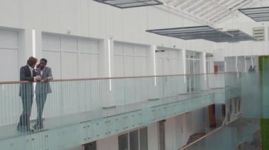 Takım elbiseli iki farklı iş adamının iş merkezindeki salonda cam parmaklıklara yaslanıp sohbet ederken geniş açılı görüntüsü.
