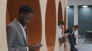 Neşeli Afrikalı iş adamının modern ortak çalışma sahasında elinde telefonla kameraya bakarken genç bir adam ve kadının arka planda telefon kullanmasını çekiyoruz.