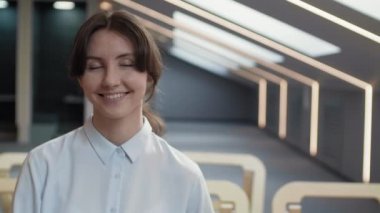 Orta ölçekli, siyah saçlı, mavi gömlekli, ahşap koltukta oturmuş kameraya bakıp gülümseyen beyaz bir kadının portresi.