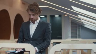 Orta boylu, yakışıklı, beyaz takım elbiseli iş adamının konferans salonunda oturmuş not defterine bir şeyler yazarken ve basın toplantısı sırasında kameraya bakarken görüntüsü.