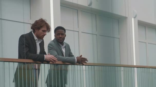 ガラスの手すりにもたれてビジネスセンターホールに立ってエレガントなスーツを着てチャットをしている若いアフリカ系アメリカ人と白人のビジネスマンの中のショット — ストック動画
