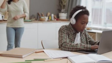 Kulaklıkla kulaklık takan, masa başında oturan, laptopunu kullanan, arka planda melez bir anneyi seven, sonra da oğlunu kontrol eden Afrikalı on yaşında bir erkek öğrencinin göğsü kalkık.