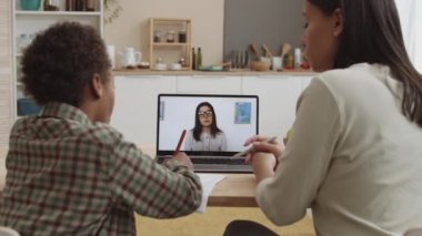 Dairede dizüstü bilgisayarın görüntüsü, genç beyaz kadın öğretmenin video dersi, annesinin kıvırcık saçlı oğluna internetten öğrenme konusunda yardım etmesi.