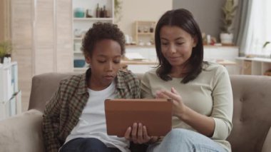 Orta boy melez bir kadının tablet bilgisayarı tutarken kanepede oturup gülümserken okul çağındaki Afrikalı oğluyla konuşurken görüntüsü.