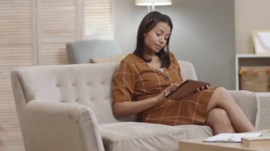 Orta uzunlukta melez bir kadın oturma odasında kanepede oturuyor, tablet bilgisayar kullanıyor, el hareketleriyle 10 yaşındaki oğlunu çağırıyor, Afrikalı çocuk nefes darlığı çekiyor, konuşuyorlar.