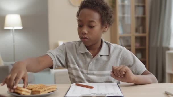 机の上に座って紙に書き宿題をしたりワッフルで軽食をしたりしましたこれはアフリカ系アメリカ人の少年が — ストック動画