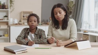 Orta ölçekli aile bireyleri evde masa başında oturuyor, okul ödevlerini birlikte yapıyor, kameraya bakıyor, arkasında görünmez biriyle konuşuyor.