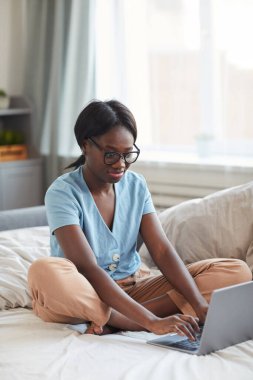 Genç Afro-Amerikan bir kadının evde yatarken dizüstü bilgisayar kullanarak dikey portresi.