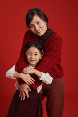 Güzel, olgun, Asyalı bir kadının, kırmızı duvarın önünde dikilip şirin küçük kızına sarılışının orta uzunlukta stüdyo resmi.
