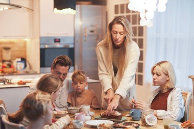 Wide angle portrait of big happy family enjoying breakfast together sitting at table in kitchen , focus on mother serving food and cutting cake, copy space