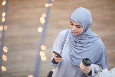 Orta Doğulu zarif bir kadının akıllı telefon kullanan ve alışveriş merkezlerinde alışveriş torbaları taşıyan yüksek açılı portresi.