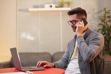Bilgisayarla çalışan ve kırmızı masada otururken telefonla konuşan yakışıklı bir adamın yan görünüm portresi.