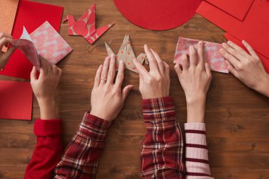 Tanımlanamayan bir kadın ve iki kızın, origami vinçleri yapmak için renkli kağıt kullanarak tahta masada oturduğu düz bir kare.