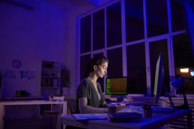 Modern genç kadın gecenin geç saatlerinde karanlık bir ofiste oturuyor kahve içiyor ve bilgisayar ekranına bakıyor.